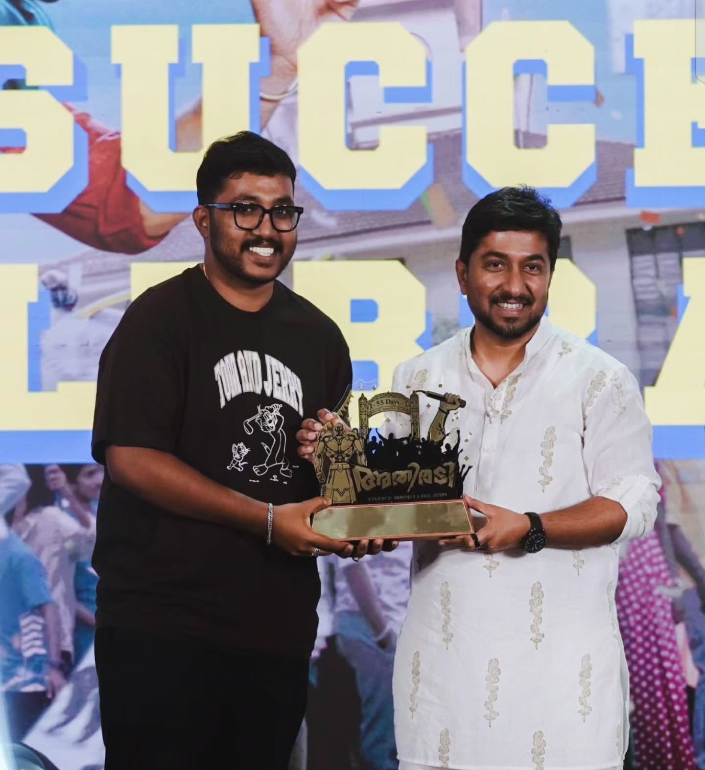 Two men standing on a stage holding a trophy. The man on the left wears a black t-shirt with a graphic print and black pants, while the man on the right wears a white kurta with gold patterns and white trousers.