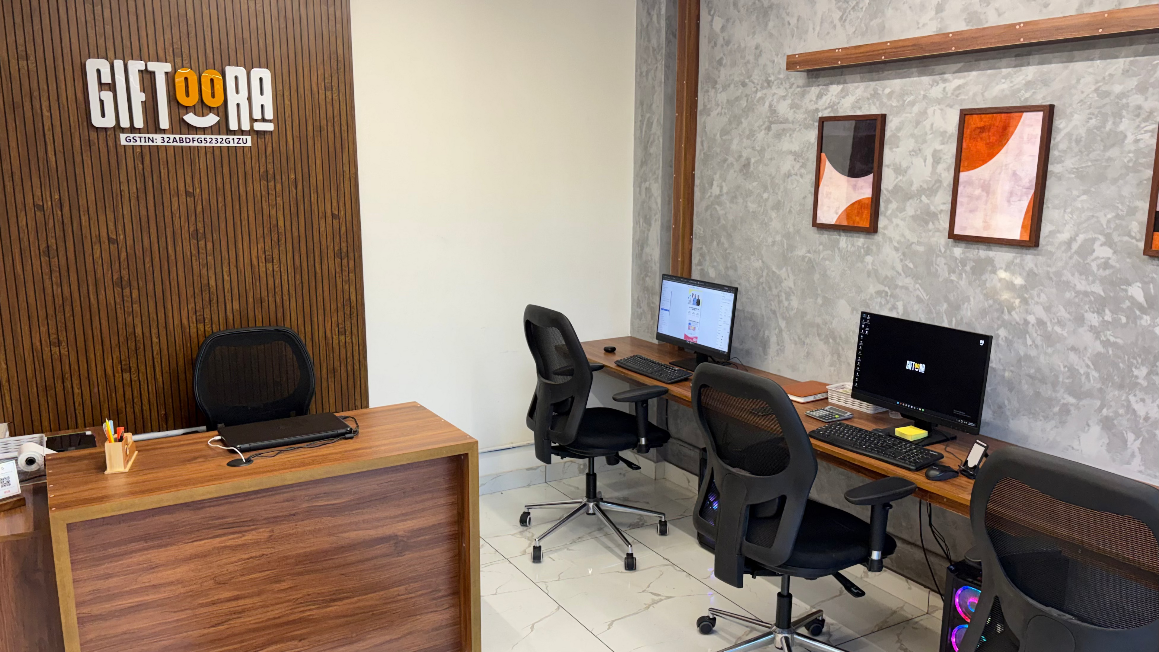 A modern office interior with a wooden reception desk on the left, a gray textured wall with two framed abstract prints on the right, and two workstations with computer monitors, keyboards, and black office chairs.
